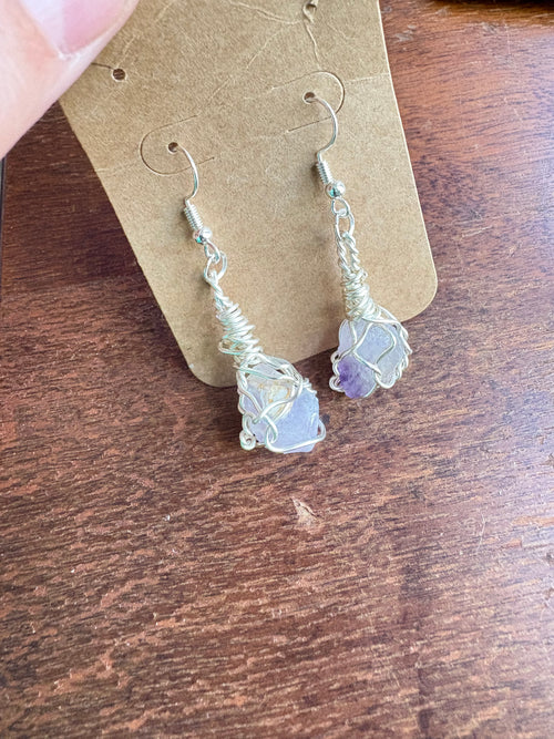 A pair of handmade wire-wrapped dangle earrings with light purple stones, displayed on a brown cardboard.