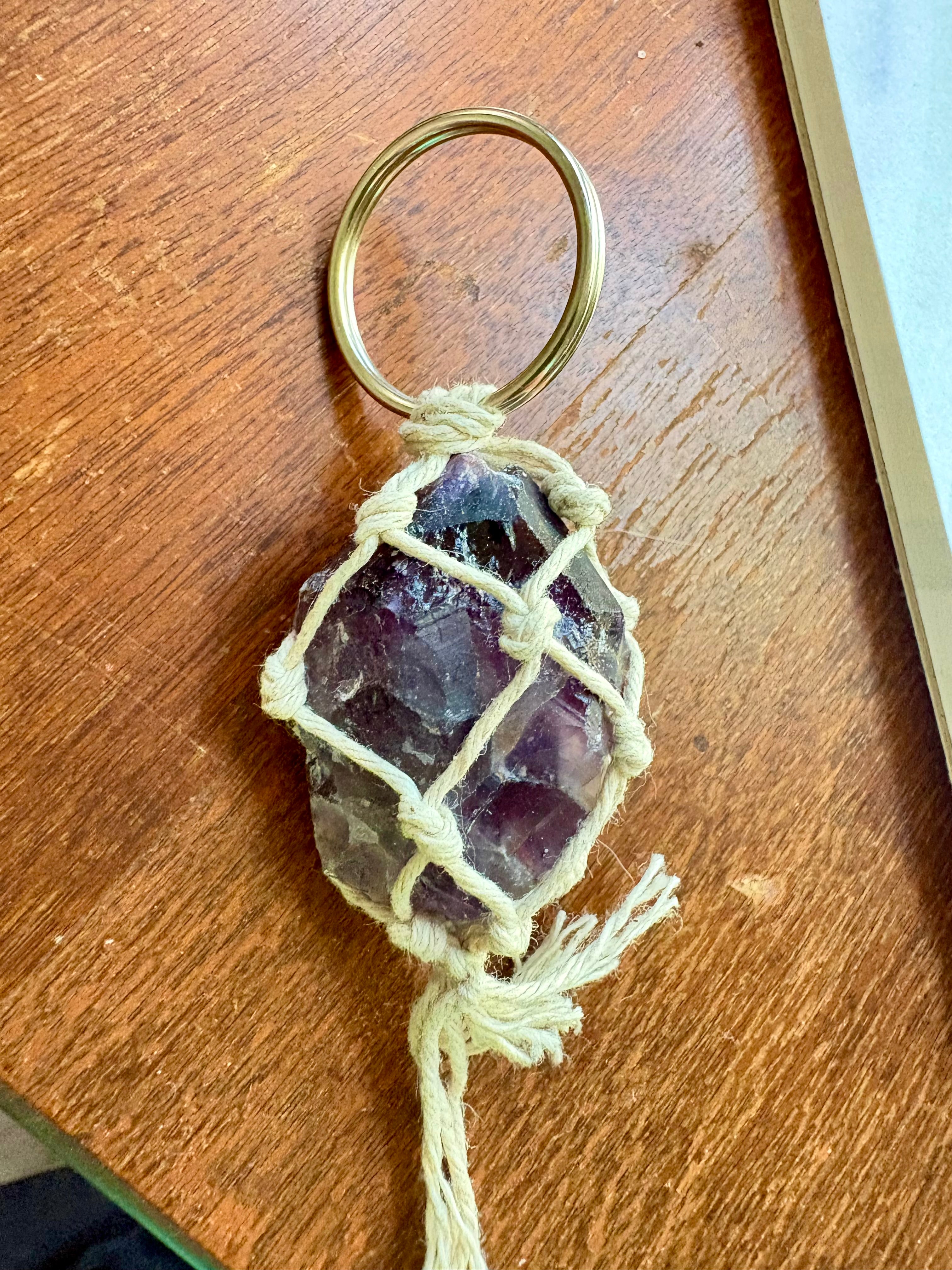 A handmade keychain featuring a raw purple amethyst crystal wrapped in cream-colored cord, attached to a silver-colored metal split ring, resting on a wooden surface.