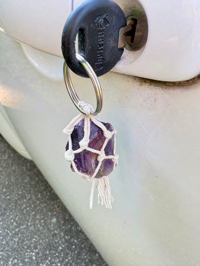 A purple amethyst crystal wrapped in a white macrame net and tassel, hanging from a metal key ring attached to a black car key fob on a white vehicle door.