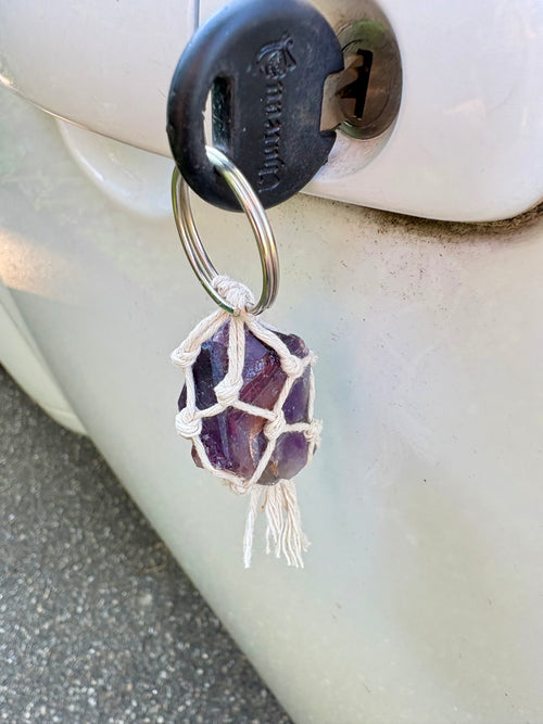 A purple amethyst crystal wrapped in a white macrame net and tassel, hanging from a metal key ring attached to a black car key fob on a white vehicle door.