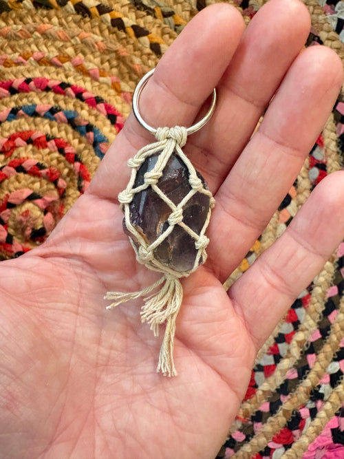 A handmade keychain featuring a raw, unpolished dark purple stone wrapped in cream-colored cord with a silver split ring