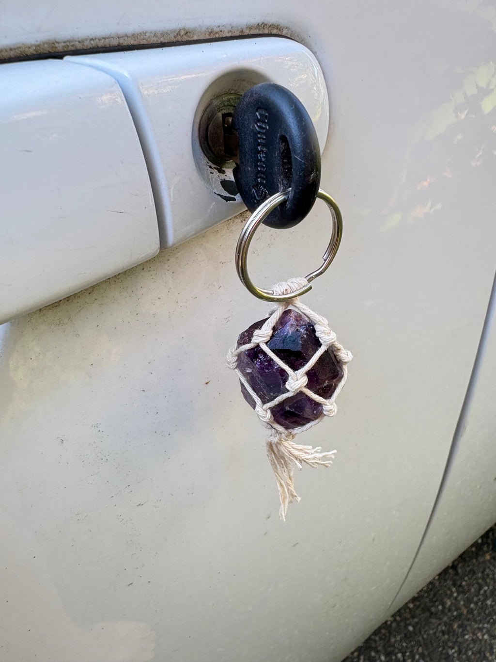 A close-up of a keychain with a black oval key fob, a silver key ring, and a purple amethyst crystal wrapped in white macrame.